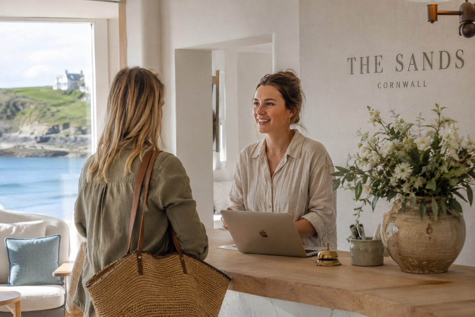 Bright, airy boutique hotel front desk in natural daylight, Cornwall coastal setting. A staff member relaxed and smiling, mid-conversation with a guest, laptop open on the counter but not the focus of the shot. Pale stone, bleached wood, soft linen tones, sea-blue accents through a window. Editorial 35mm style, candid, not posed. No screens showing obvious "AI" branding, no robots, no dark shadows.